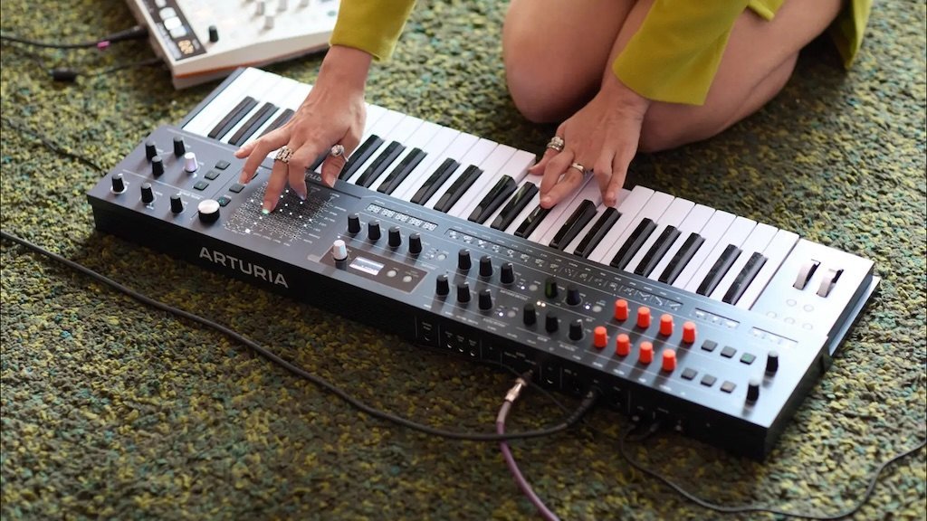 Two hands playing the Arturia MiniFreak 49 synthesizer on a green rug.