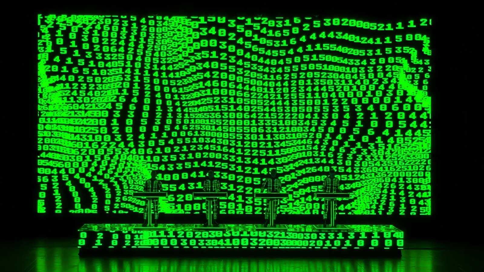 Kraftwerk on stage: four performers in grid-patterned suits at lit podiums before a giant screen of green scrolling numbers.