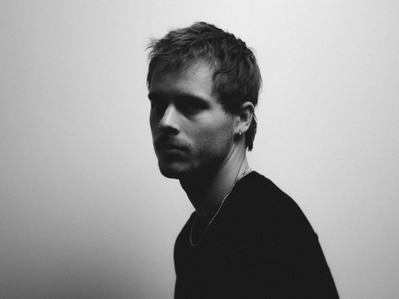 Black-and-white portrait of Riordan in a dark top and chain necklace, head turned to the side against a plain light background