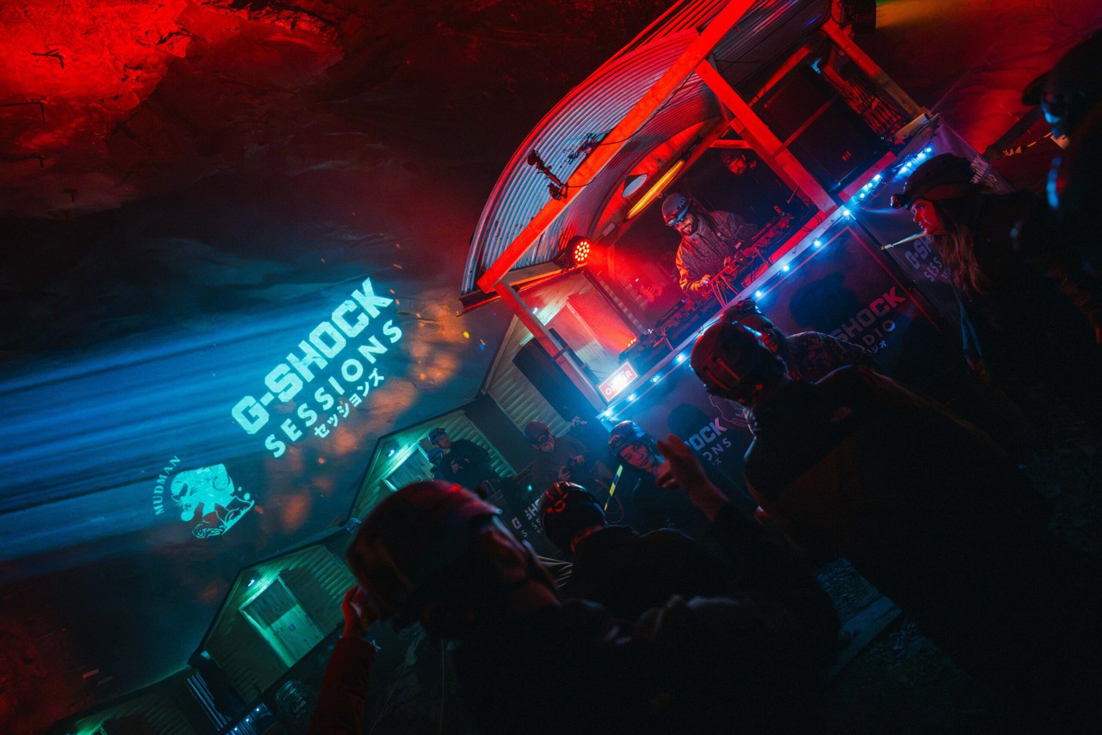 DJ at the decks in a lit booth inside a dark cave chamber, with a "G-SHOCK Sessions" and Mudman logo projected onto the rock and a crowd in helmets in the foreground